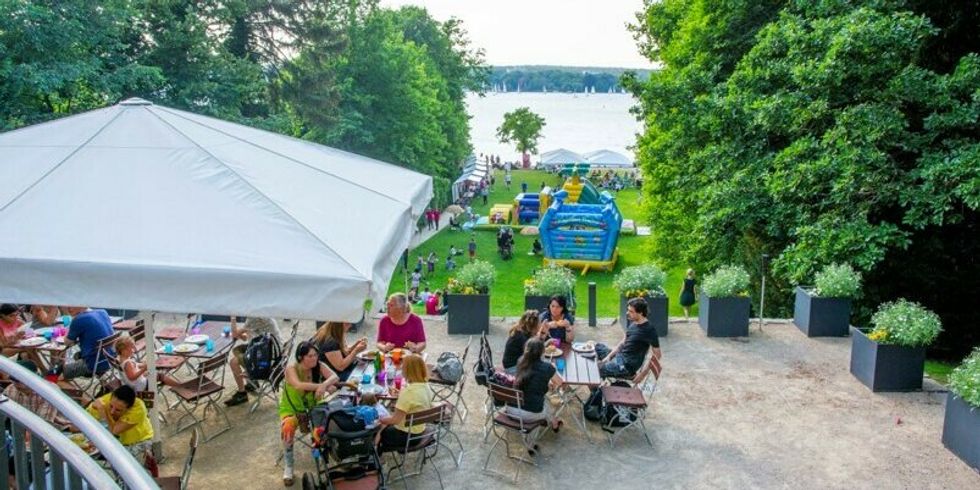 Menschen sitzen beisammen am Wannsee und genießen das schöne Wetter. Im Hintergrund ist eine blaue Hüpfburg und der Wannsee zu sehen.