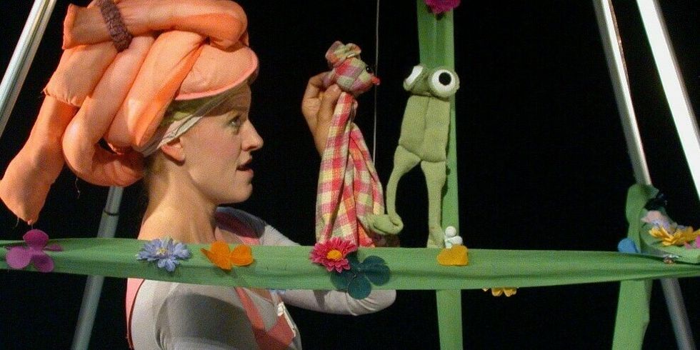 Eine bunt gekleidete Frau steht zwischen einem Gestellt mit Puppen und hält eine in der Hand.