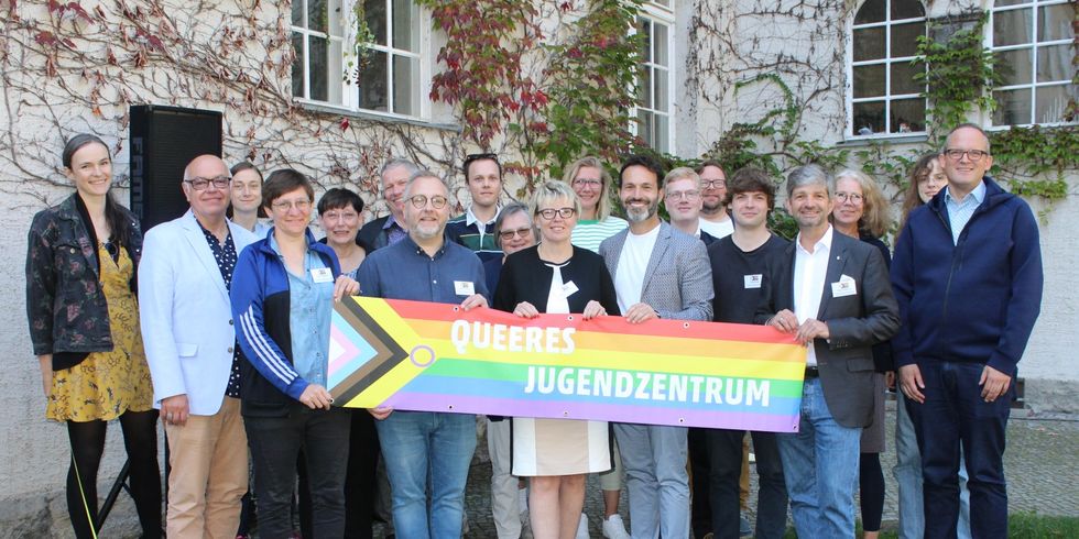 Mehrere Personen stehen zusammen vor einem Haus und schauen in die Kamera. Sie halten eine große LGBTIQ Flagge