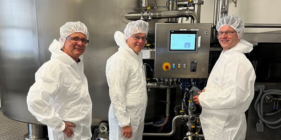 Drei Männer stehen in sterilen weißen Anzügen neben einer Maschine in einer Produktionshalle.