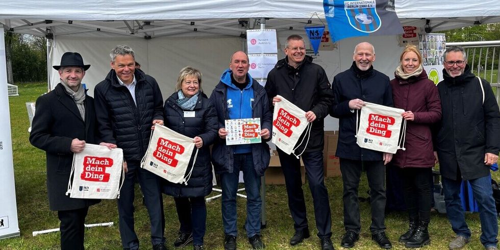 Mehrere Personen stehen auf einem Feld vor einem Zelt. Es werden Beutel und ein Flyer hochgehalten mit der Aufschrift "Mach dein Ding".