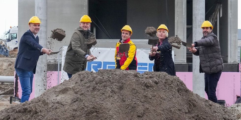 Fünf Personen mit gelben Schutzhelmen schippen Erde mit Spaten auf einen Haufen.