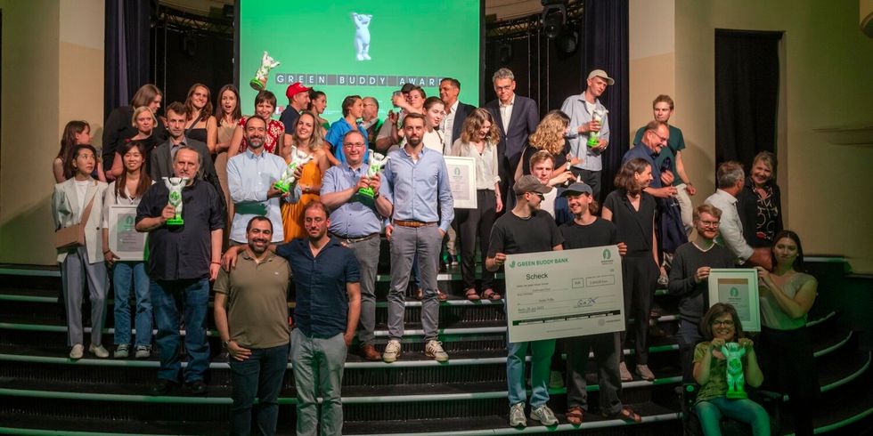 Eine Gruppe Menschen mit Preisen, Urkunden und Schecks steht auf einer Treppe vor einer Bühne. Im Hintergrund ist eine Leinwand mit dem Schriftzug "Green Buddy Award".