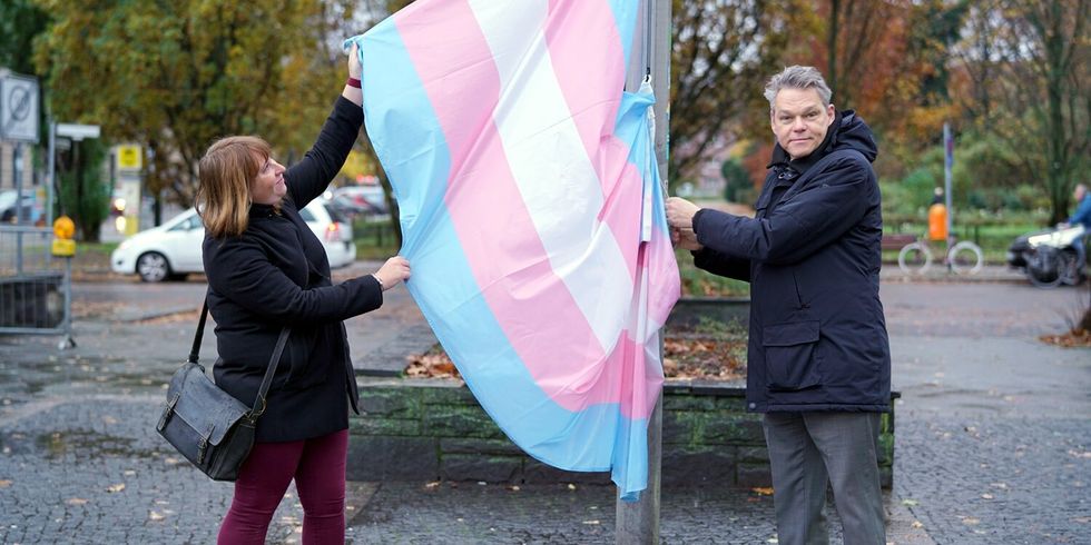 Eine Frau und ein Mann stehen auf einem Platz und halten eine blau-rosa-weiße Fahne an einem Fahnenmast.