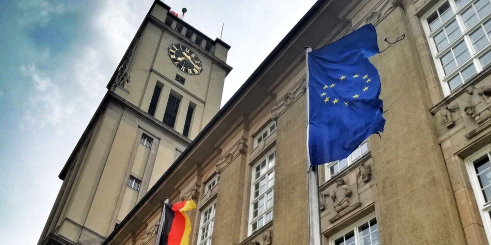 Vor einem großen Gebäude mit Uhrenturm wehen die deutsche Flagge und die EU-Flagge.