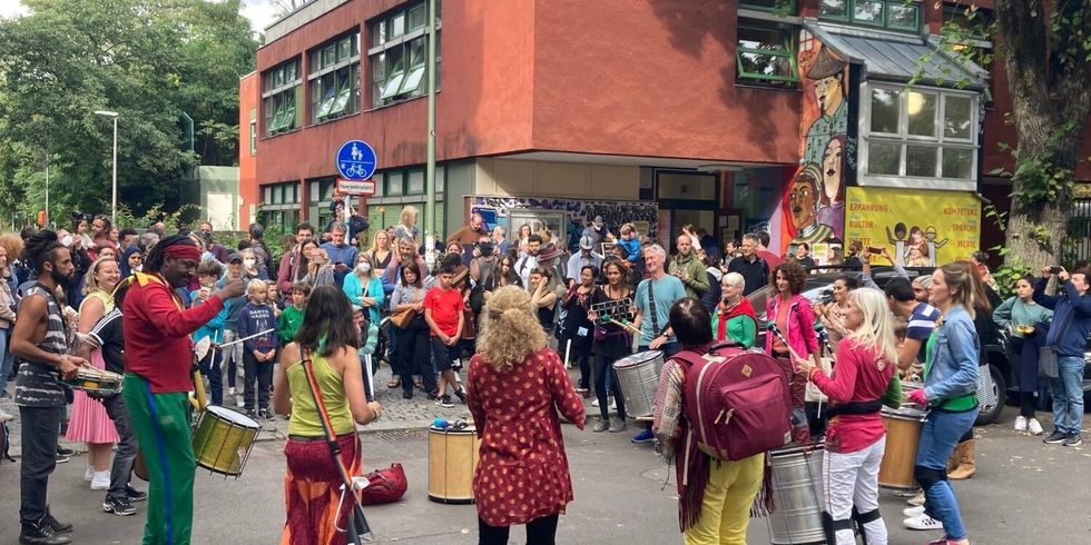 Auf einer freien Fläche vor einem Haus stehen Menschen in einem Kreis. In der Mitte wird mit Trommeln Musik gemacht.