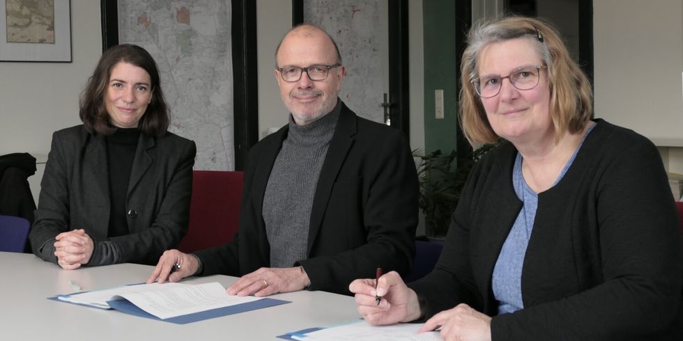 Zwei Frauen und ein Mann sitzen nebeneinander an einem Tisch. Eine Frau hat einen Stift in der hand und ist dabei etwas zu unterschreiben.