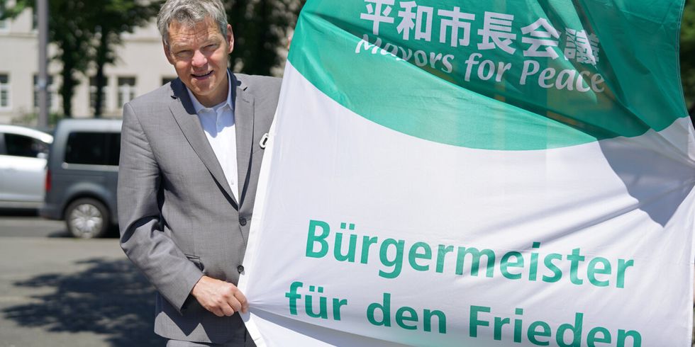 Ein Mann im Anzug hält eine grün-weiße Flagge mit der Aufschrift "Bürgermeister für den Frieden".