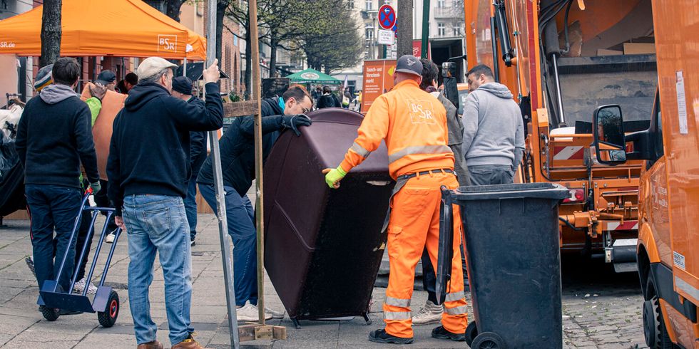 An einer Straße packen Menschen, darunter ein Mann mit orangener Arbeitskleidung der BSR, einen Schrank und versuchen ihn zu heben. Am Straßenrand stehen mehrere Wägen der BSR.