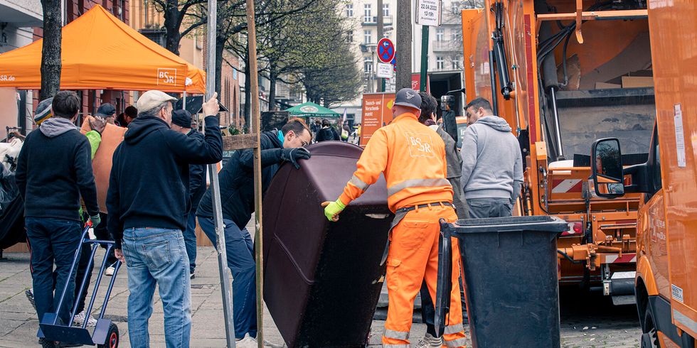 Auf einem Gehweg tragen zwei Männer eine Couch. Einer der Männer trägt die Uniform der BSR. Am Straßenrand sind BSR-Wägen abgestellt und im Hintergrund sind weiter Menschen zu erkennen, einer zieht einen Handwagen hinter sich her.