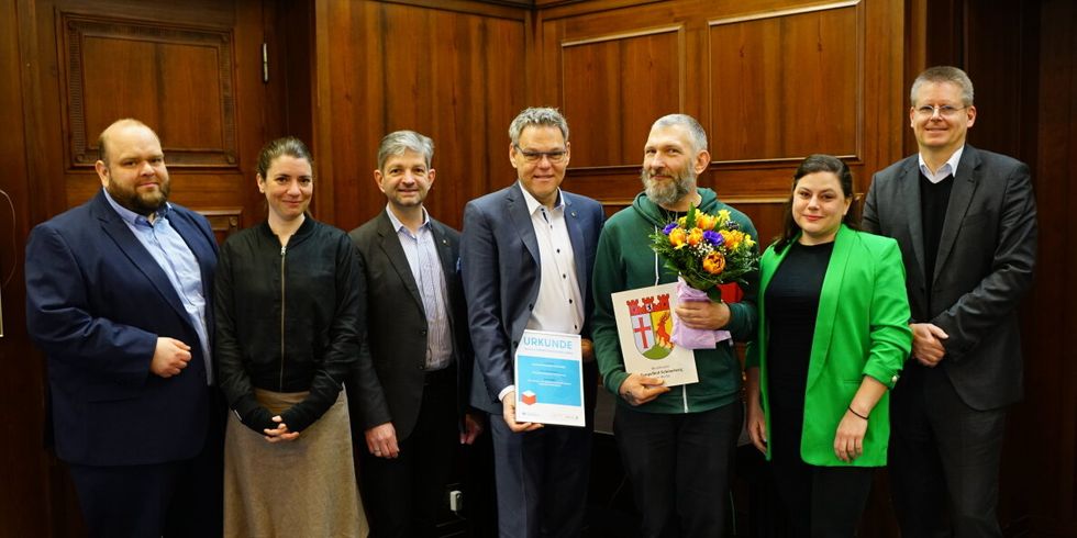 Sieben Personen stehen nebeneinander in einem Saal. Ein Mann hält einen Blumenstrauß und eine Mappe mit dem Wappen von Tempelhof-Schöneberg in den Händen. Ein weiterer Mann hält eine Urkunde.