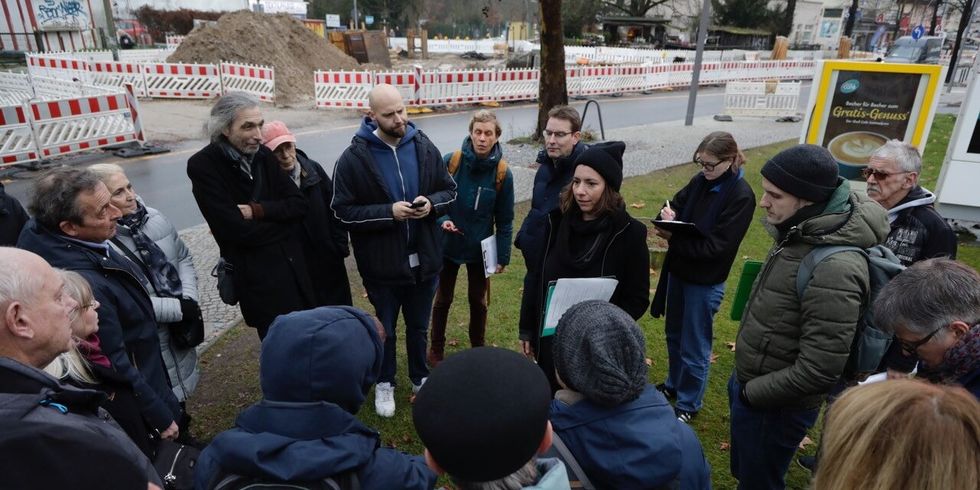 eine Frau spricht umgeben von einer kleinen Gruppe von Personen vor einer Baustelle