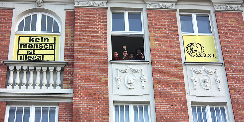 Fenster an einer Hausfassade. An zwei Fenstern sind Banner. Auf einem Banner steht "Kein Mensch ist illegal" und auf dem anderen steht "SUSI" und ein Logo. Aus einem Fenster in der Mitte schauen Frauen heraus und winken.