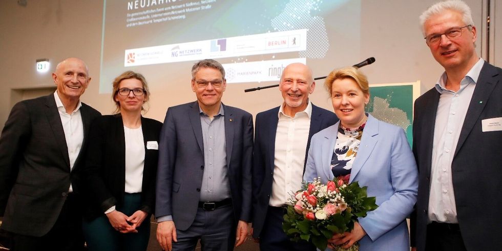 Sechs Personen stehen nebeneinander und lächeln in die Kamera. Eine Frau hält einen Blumenstrauß.