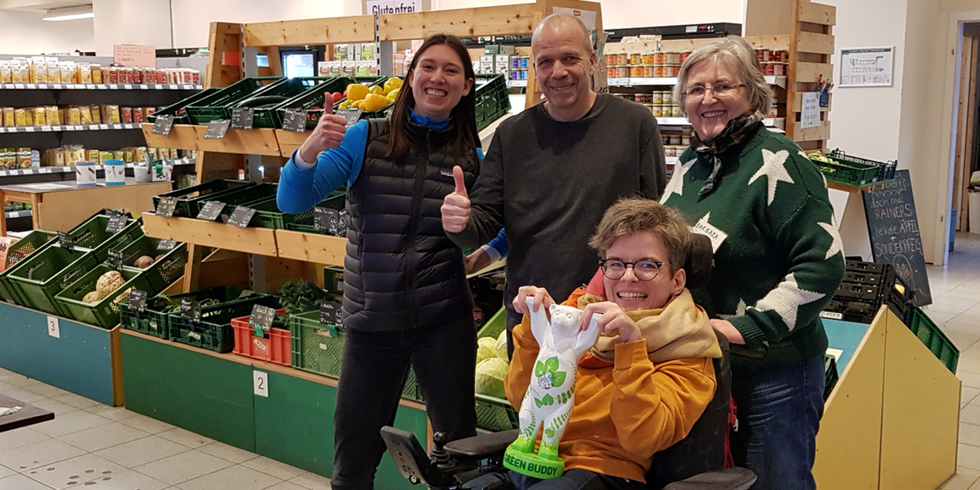 In einem Supermarkt stehen bzw. sitzen vier Personen und lächeln in die Kamera. Zwei Personen zeigen einen Daumen nach oben und eine Person im Rollstuhl hält eine Figur eines Bären mit der Aufschrift "Green Buddy" in den Händen.