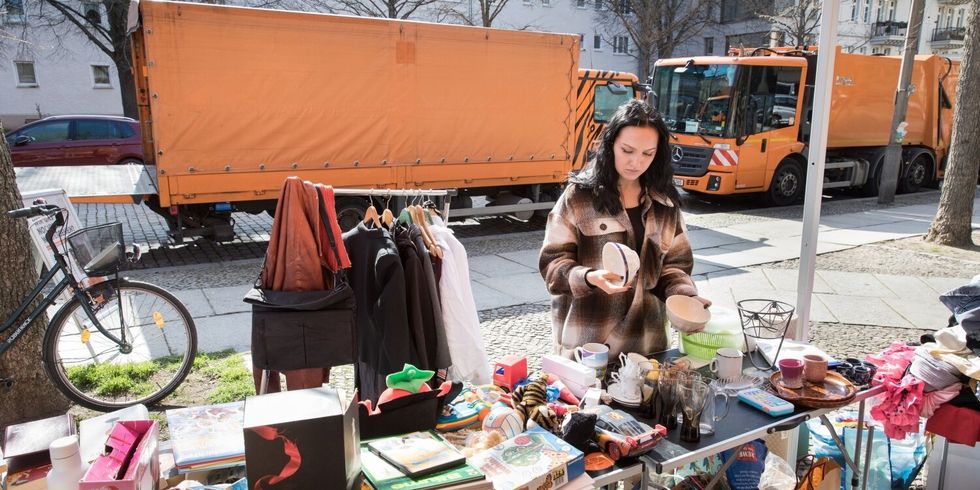 Eine Frau steht an einem Straßenrand an einem Stand mit unterschiedlichen Dingen. Hinter ihr stehen zwei große Wägen der Berliner Stadtreinigung.