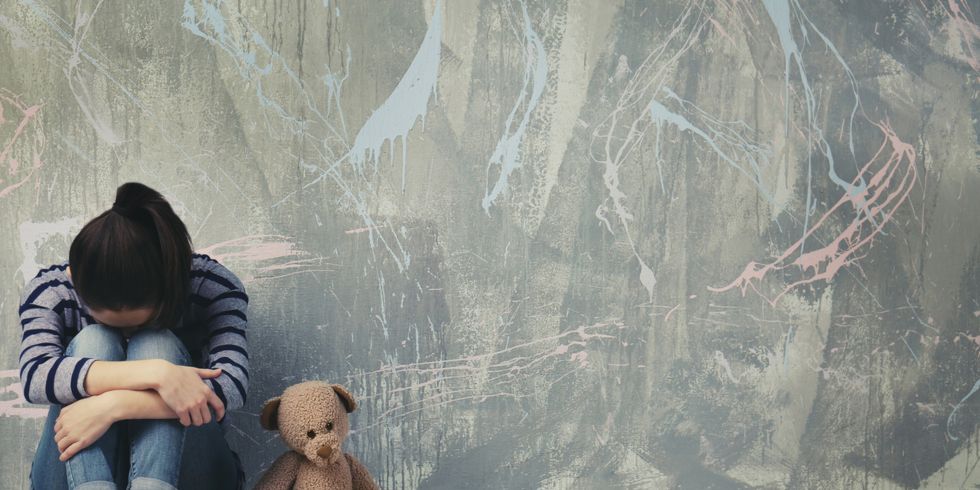 Eine junge Frau sitzt mit angezogenen Knien am Boden, daneben ein Teddybär.