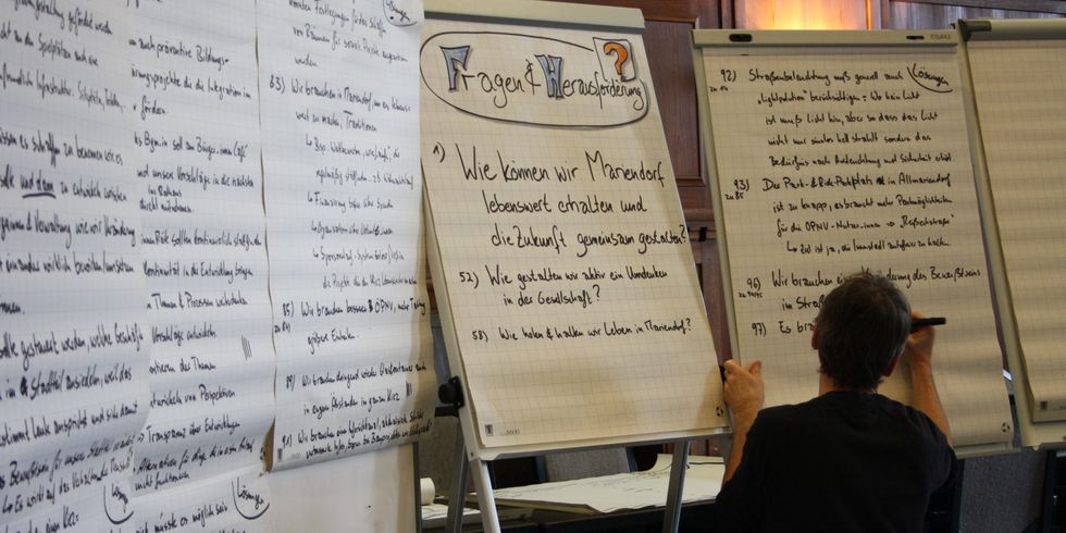 Mehrere eng beschrieben Flipchart hängen nebeneinander, eine Person schreibt auf eines von ihnen.