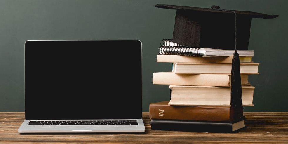 Laptop und Bücherstapel auf einem Tisch, akademische Mütze liegt auf dem Bücherstapel