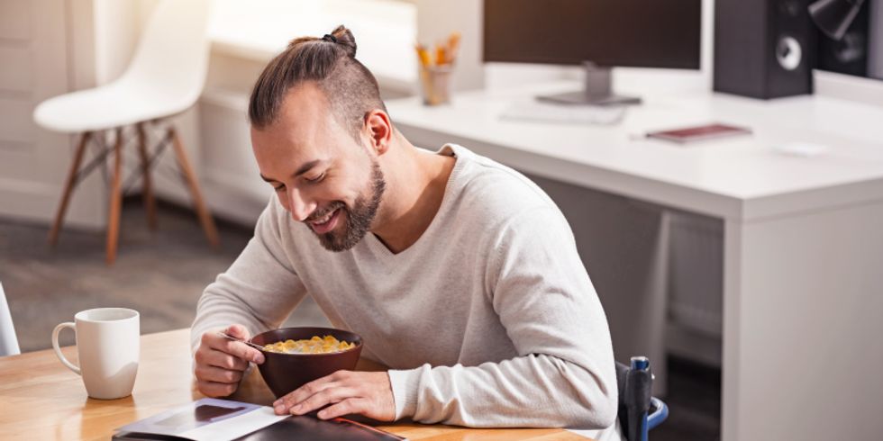 Junger Mann im Rollstuhl genießt seinen Kaffee und das Müsli am Morgen, während er beiläufig in einem Buch auf seinem Tisch liest