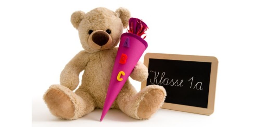 Teddybär mit einer Schultüte mit der Aufschrift "ABC" und einer Tafel auf der "Klasse 1a" steht