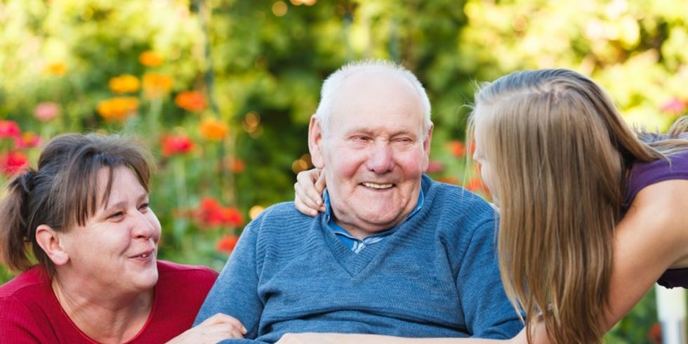 Senior im Rollstuhl wird von seiner Tochter und Enkelin umarmt. Alle drei lächeln sich an.