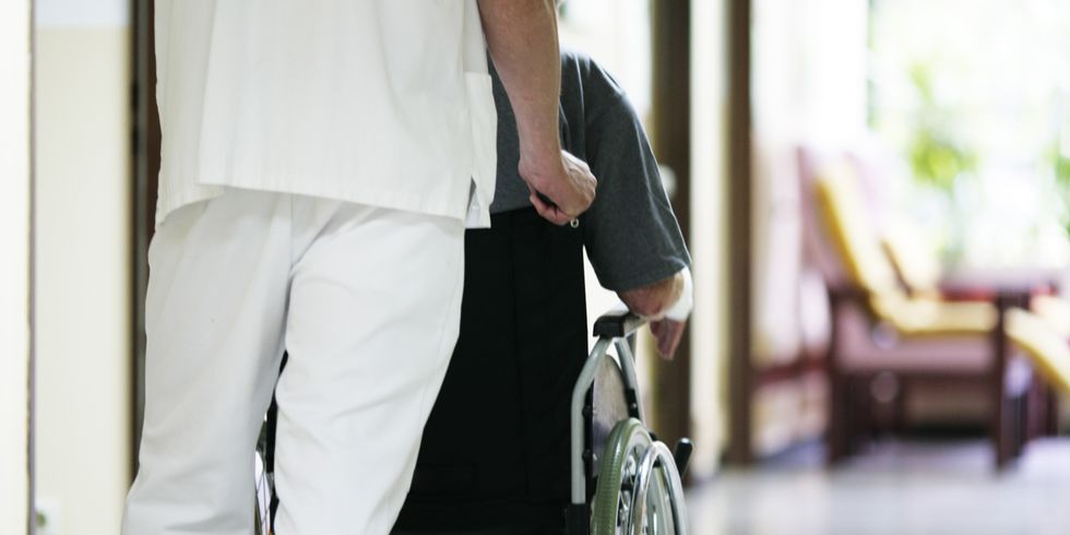 Auf der Pflegestation schiebt ein Pfleger einen Menschen mit Behinderung im Rollstuhl.