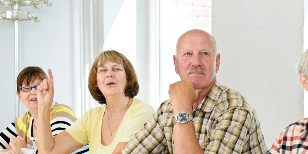 Senioren sitzen an einem Tisch mit Büchern, eine Seniorin hebt den Zeigefinger und setzt zum Sprechen an