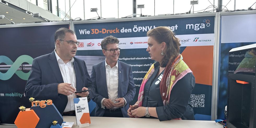 3D-Druck-Präsentation auf einer Messe. Drei Personen stehen vor einem Tisch mit 3D-Druck-Modellen und einem Banner mit dem Titel „Wie 3D-Druck den ÖPNV bewegt"