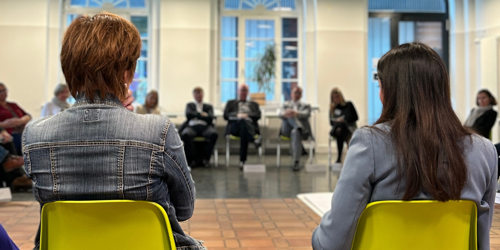 Das Bild zeigt zwei Frauen von hinten, sie sitzen auf grünen Stühlen und blicken auf eine Reihe von Menschen, die man unscharf im Hintergrund sieht.