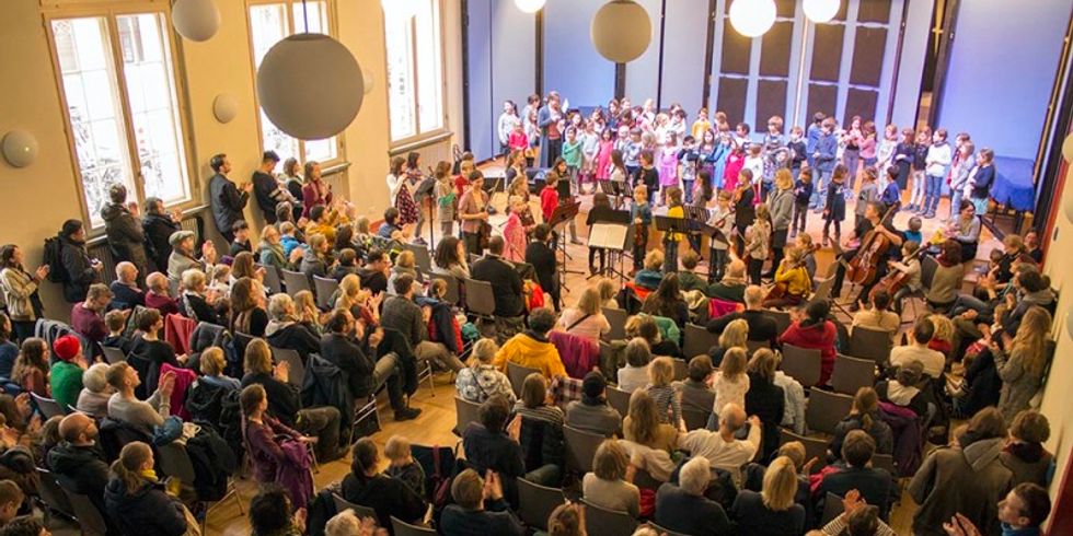 Voller Konzertsaal mit Bühne, darauf singende und musizierende Kinder