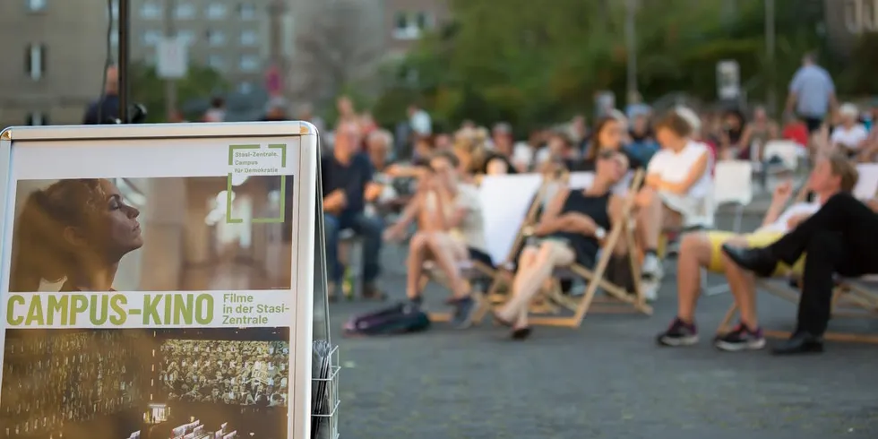 Vordergrund: Werbeaufsteller "Campus-Kino" Hintergrund: Kino-Publikum in Sonnenstühlen