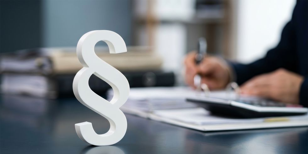 Paragraphensymbol steht auf einem Tisch, dahinter arbeitet eine Person an Papierunterlagen