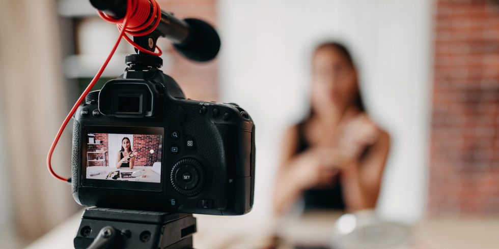 Im Vodergrund eine Kamera auf einem Stativ aufgebaut, welche eine Frau bei einem Video filmt