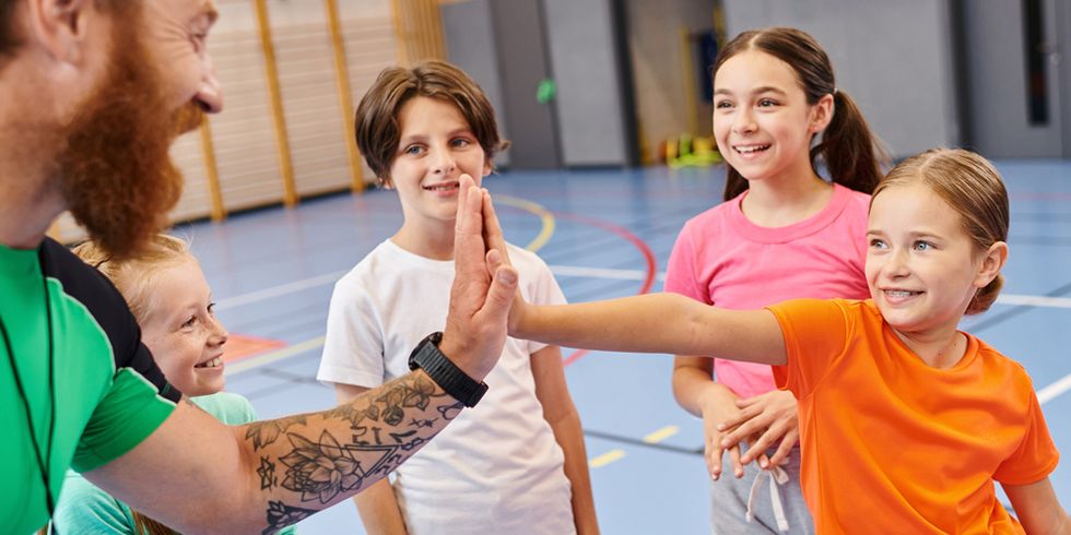 Gruppe junger Kinder steht begeistert in einer Turnhalle und klatschen mit dem Lehrer ein