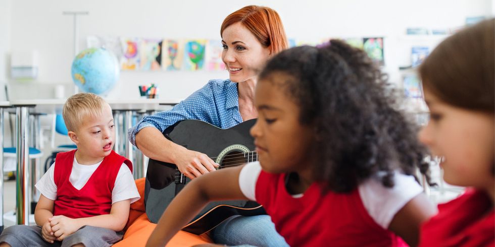 Schulkinder und Lehrerin mit Gitarre sitzen im Klassenzimmer, ein Kind mit geistiger Behindung