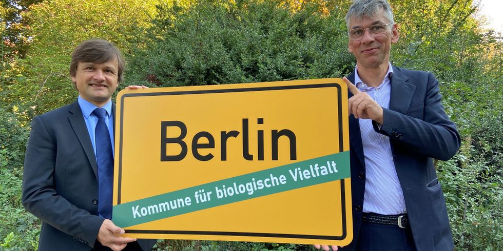 Vor grünen Büschen halten Robert Spreter und Staatssekretär Stefan Tidow in Hemd und Jackett das gelbe Berliner Ortseingangsschild hoch. Unter dem Stadtnamen steht: Kommune für biologische Vielfalt. Anlass ist der Beitritt Berlins dem Bündnis am 1. Oktober 2020.