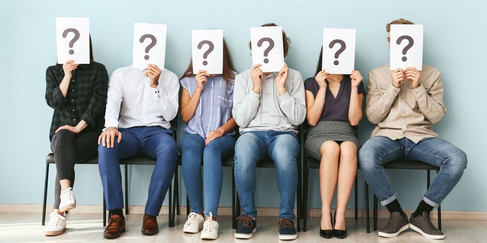 6 Menschen sitzen in einer Reihe und halten sich ein Blatt mit Fragezeichen vor das Gesicht