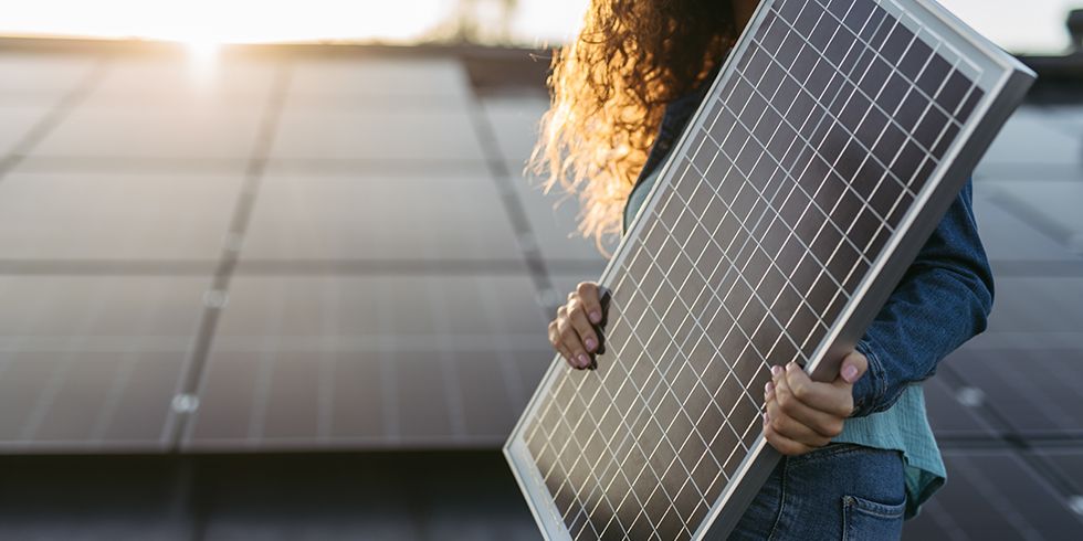 Frau hält Solarpanel in den Händen. Im Hintergrund Solarmodule auf Dach.
