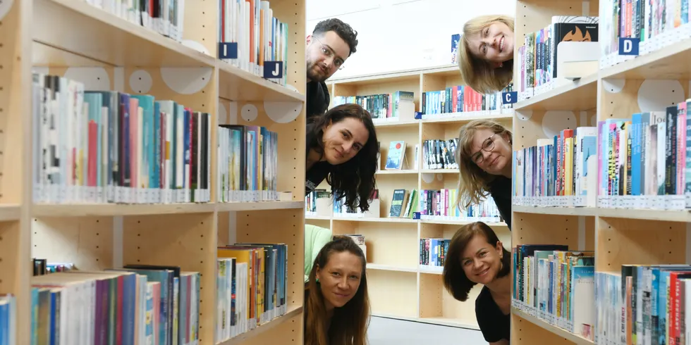Sechs Bibliothekar:innen verstecken sich hinter den Bücherregalen.