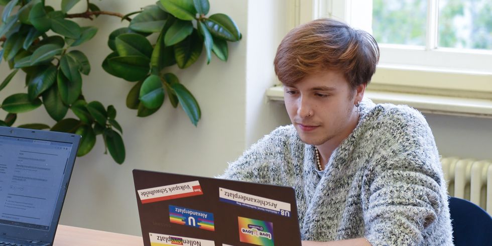 Junge Erwachsene mit einem Laptop in der Bibliothek. Auf dem Laptop sind viele Queer-Pride-Aufkleber.