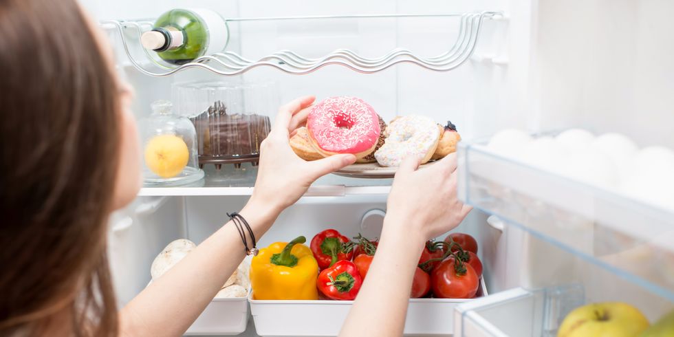 Ein offener Kühlscrhank mit Gemüse und Gebäck. Eine Frau nimmt sich Donuts.