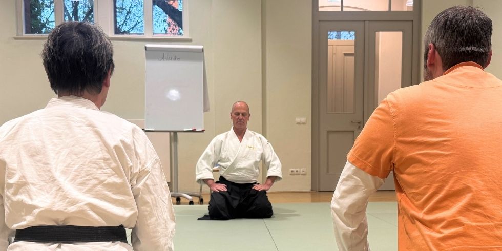 Kursleiter Klimpel und zwei Teilnehmer in der Turnhalle sitzen sich im Fersensitz gegenüber