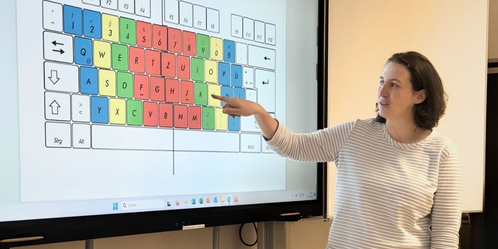 Kursleiterin Neig steht vor dem Whiteboard. Sie zeigt auf eine abgebildete Tastatur. Die Tasten sind unterschiedlich farbig.
