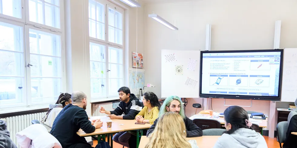 Teilnehmende eines Deutschkurses sitzen an Gruppentischen in einem Unterrichtsraum vor einem interaktiven Whiteboard.