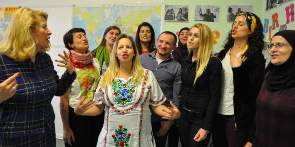Eine Gruppe von Erwachsenen steht in einem Klassenzimmer und singt oder spricht gemeinsam. Im Vordergrund trägt eine Frau ein traditionell besticktes weißes Kleid. Im Hintergrund sind eine Weltkarte, Fotos und bunte Buchstaben an der Wand zu sehen.