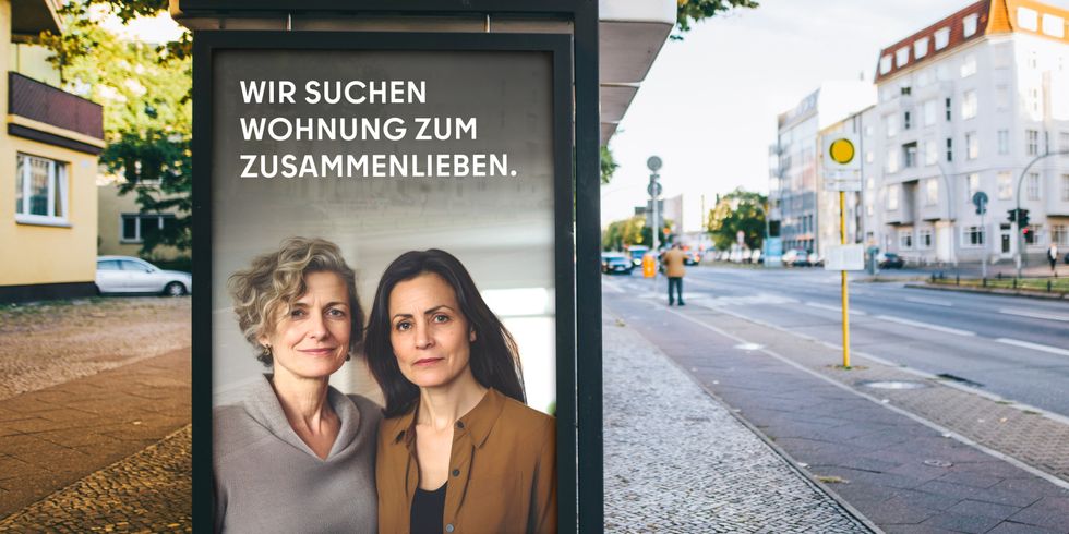 Das Bild zeigt eine Werbetafel an einer Straße. Auf der Tafel sind zwei Frauen abgebildet, die nebeneinander stehen und in die Kamera blicken. Der Text auf der Tafel lautet: “WIR SUCHEN WOHNUNG ZUM ZUSAMMENLIEBEN.” Im Hintergrund sind Gebäude, eine Straße und ein Fußgänger zu erkennen.