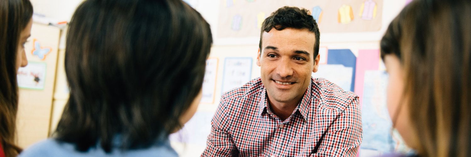 Ein Lehrer sitzt drei Schülerinnen gegenüber und spricht