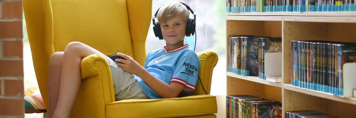 Ein Junge sitzt im Sessel in einer Bibliothek und hat Kopfhörer auf dem Kopf und ein iPad in der Hand