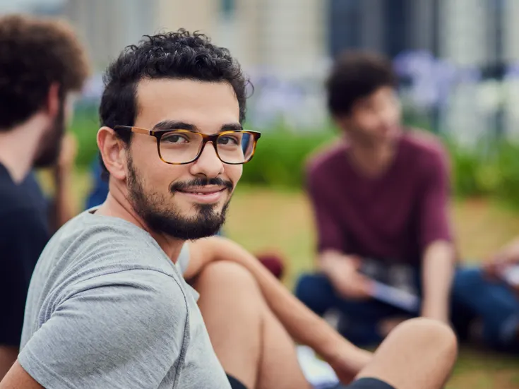 Ein Portrait eines junges Mannes mit Brille, der in eine Gruppe von jungen Menschen sitzt.